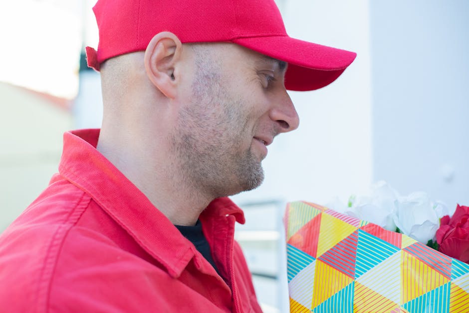 A man dressed in a red cap and matching red jacket is holding a vibrant floral gift wrapped in colorful, geometric-patterned paper. The flowers inside the wrapping include pink and white blooms, with 