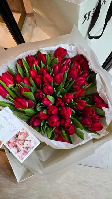 A large bouquet of fresh red tulips with vibrant green stems and leaves, arranged in a rounded cluster. The tulips are wrapped in white paper with a slightly crinkled texture, lined with tissue paper.