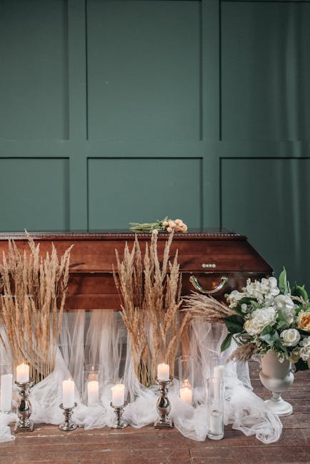 A floral arrangement featuring white roses and green foliage in a white vase, placed on a table draped with white tulle fabric. Surrounding the vase are tall candles in silver holders and decorative p