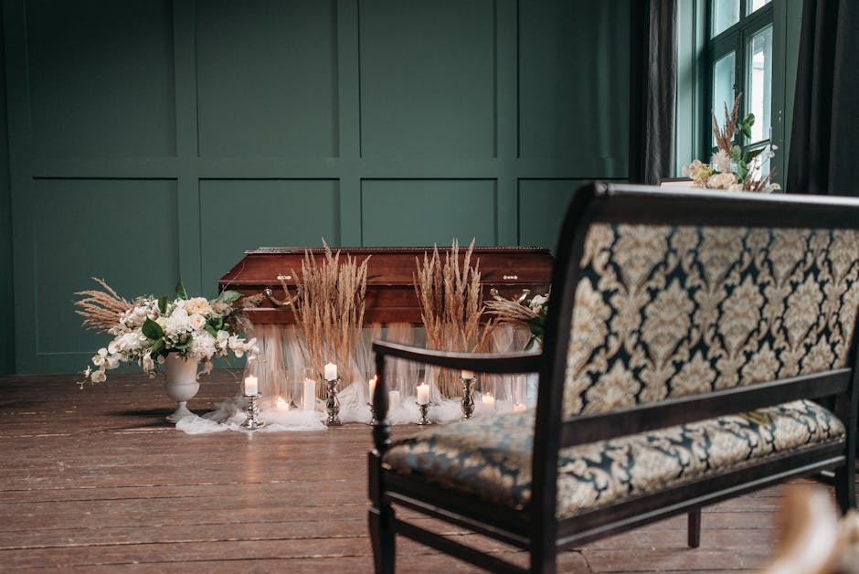A floral arrangement on a wooden table featuring tall, beige pampas grass stems, complemented by white roses and other delicate white flowers in a white vase, arranged as a sympathy floral tribute. Sm