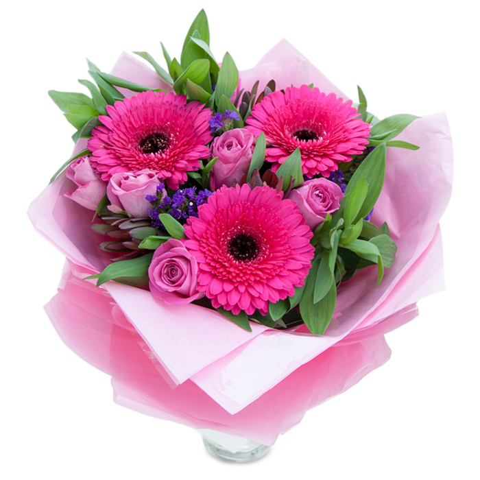 This bouquet reads as an intimate celebration in pinks and purples: three bold fuchsia Gerbera daisies open wide with radiating petals and dark, central disks that create a dramatic focal point. Interlaced among them are roses in a dusty-lavender pink, their petals still held in a fresh, sculpted curl that suggests newness and tenderness. Sprigs of purple statice are tucked into the spaces between the major blooms, their tiny clustered florets introducing a delicate, papery texture and a cool tonal counterpoint. A generous layering of deep green foliage - broad leaves that resemble salal or eucalyptus - provides weight and movement, forming a verdant collar that accentuates the bouquet's shape. The floristry presentation includes an inner wrap of pale pink, matte tissue paper and an outer veil of translucent fuchsia-pink cellophane, slightly crinkled to catch the light and echo the vivid tones of the flowers; the wrapping is folded with practiced hands, signalling careful preparation. The lower section reveals the lip of a clear glass vase, suggesting the arrangement is staged for prompt display on a mantle or as a dining table centrepiece. Sense-wise, the composition offers a soft rose fragrance rounded by a hint of green from the gerberas, and the textural contrast between smooth petals, papery statice, and glossy foliage invites touch. For residents in Creekmouth and surrounding Barking neighbourhoods, this bouquet feels suited to an affectionate birthday greeting, a thoughtful anniversary surprise, or a cheerful 'just because' delivery that lifts a room with fresh colour and earnest craft.
