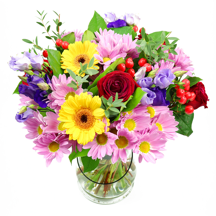 The photograph shows a generous, mixed-colour bouquet arranged in a clear glass vase, positioned against a crisp white background that enhances the vivid contrast of each bloom. Dominating the centre are several deep red roses, their velvety petals spiralling inward to create a sense of richness and emotional depth. Nestled alongside these are two bright yellow gerbera daisies, each with a dark brown-orange centre that adds a warm, earthy accent at the heart of their radiant petals. These bold tones are softened by a halo of soft pink daisies or chrysanthemums that encircle the central flowers; their many fine petals and small yellow centres give the bouquet a gentle, almost cloud-like volume. Threading their way among the pinks are graceful purple and lavender lisianthus, their bell-shaped blooms nodding slightly and adding cool, sophisticated notes, reminiscent of evening light along the Creekmouth riverside. Small clusters of glossy red hypericum berries peek through at intervals, like tiny jewels hidden in foliage, introducing rounded forms and a playful, tactile contrast to the flatter petals. A variety of green foliage frames and supports the flowers-broad, darker leaves provide structure and grounding, while slender, lighter green sprigs add freshness and a touch of movement. The clear vase at the base reveals an orderly fan of stems immersed in clear water, hinting at the professional care behind the arrangement. Overall, the bouquet conveys joy, warmth, and celebration, ideal for sending congratulations, brightening a new home, or delivering a thoughtful surprise to someone in Creekmouth, perhaps in one of the residential streets close to the Thames or near the local business parks that define the neighbourhood's character.